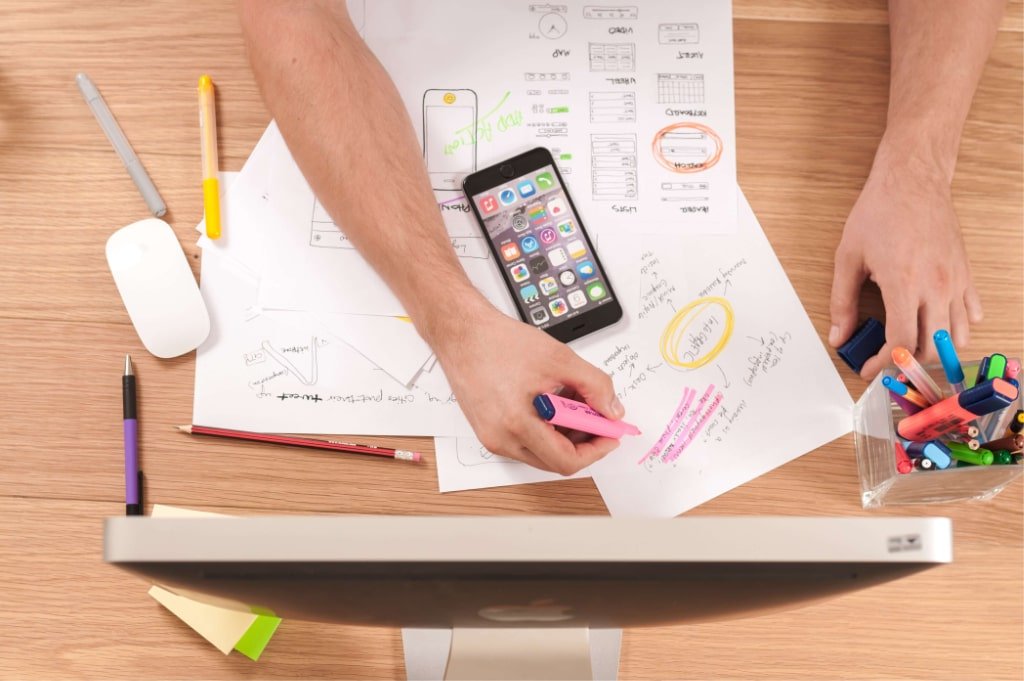 Overhead view of hands sketching a project plan on paper at a desk, with a smartphone, monitor, and stationery nearby